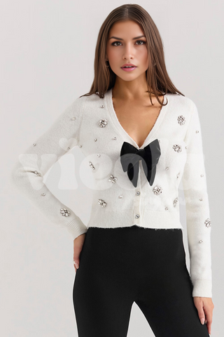 Woman wearing a white embellished cardigan with a black bow and black pants on a light gray background